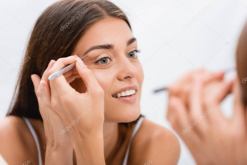 cheerful young woman tweezing eyebrows near mirror, blurred foreground