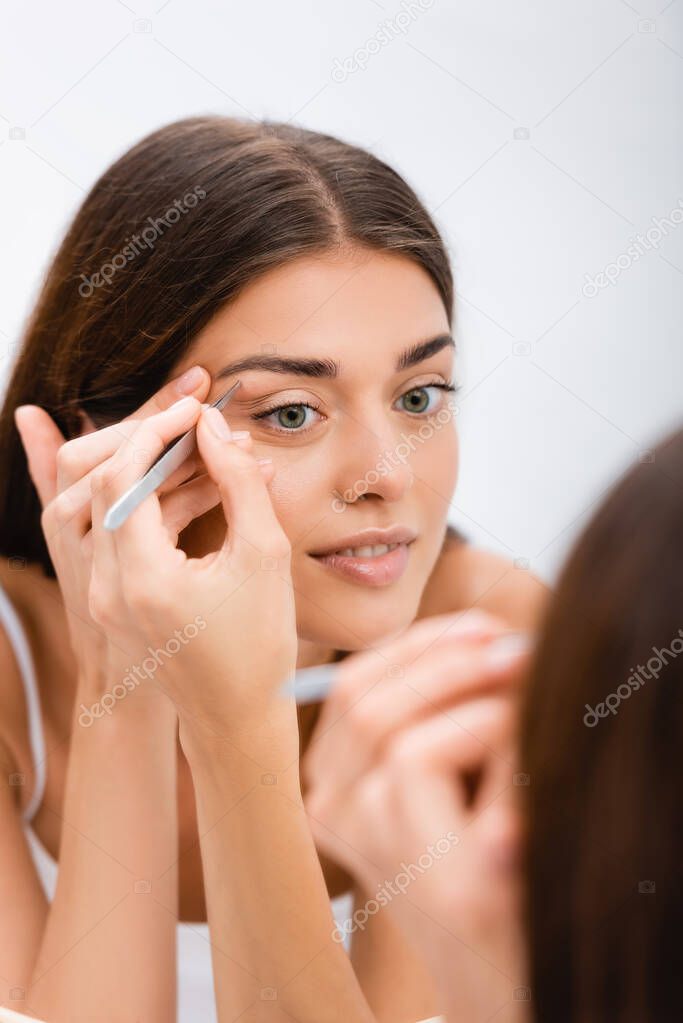 young woman tweezing eyebrows near mirror, blurred foreground