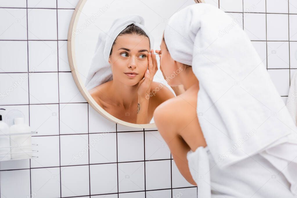 young woman with white towel on head tweezing eyebrows near mirror in bathroom