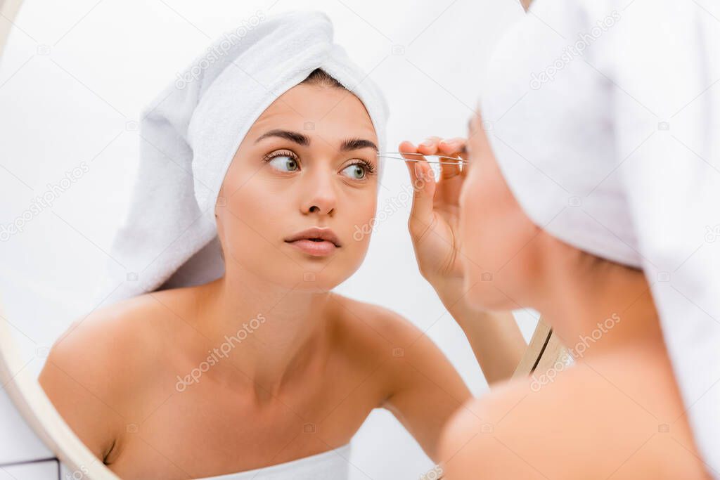 woman with white towel on head tweezing eyebrows in bathroom near mirror, blurred foreground