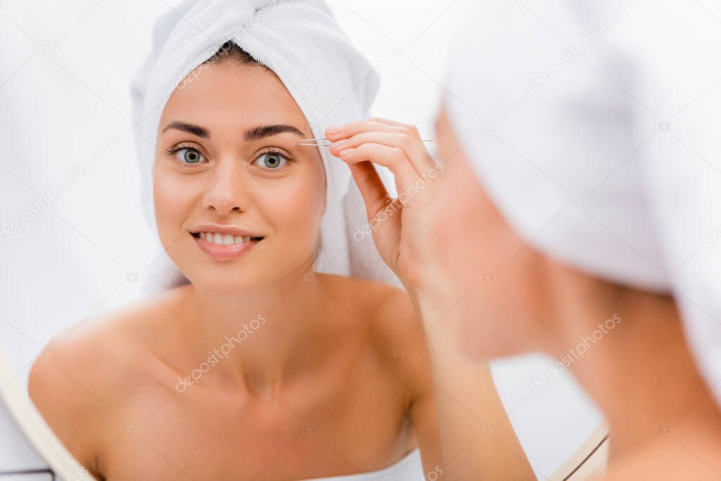 happy woman with white terry towel on head tweezing eyebrows in bathroom, blurred foreground