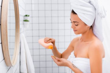 Yüzünde beyaz havluyla gülümseyen kadın banyoda yüz fırçasıyla şişeyi tutuyor.