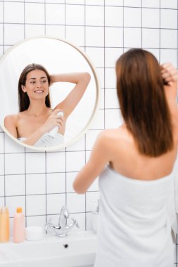 Gülümseyen kadın, beyaz havluya sarılı, banyoda aynanın yanında deodorant kullanıyor, bulanık ön plan