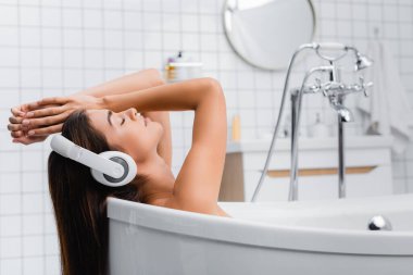 Genç bir kadın gözleri kapalı banyo yapıyor ve kablosuz kulaklıkla müzik dinliyor.