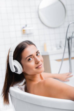 Mutlu genç bir kadın banyo yaparken kameraya bakıyor ve kablosuz kulaklıkla müzik dinliyor.