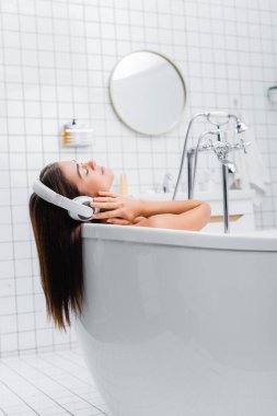 Genç bir kadın banyo yapıyor ve kablosuz kulaklıkla gözleri kapalı müzik dinliyor.