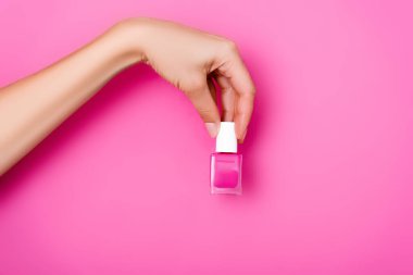 Kırpılmış bir manzara, elinde pembe arka planda parlak tırnak cilası şişesi tutan bir kadın.