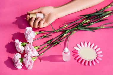 cropped view of woman holding pastel nail polish near carnation flowers, cuticle remover and palette of artificial nails on pink background clipart