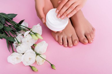 Çıplak ayaklı bir kadının pembe arka planda beyaz Eustoma çiçeklerinin yanında kozmetik krem tutarken kısmi görüntüsü