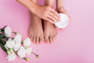 Çıplak ayaklı bir kadının pembe arka planda beyaz Eustoma çiçeklerinin yanında kozmetik krem tutuşu.