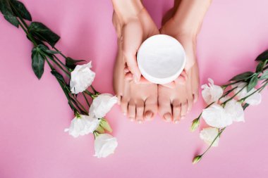 top view of female hands with cosmetic cream near feet and white eustoma flowers on pink background clipart