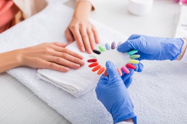 partial view of manicurist showing palette of fake nails to client on blurred background clipart