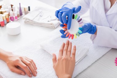 Manikürcünün, müşterinin yanındaki sahte tırnak paletini parmağıyla göstermesinin kısmi görüntüsü