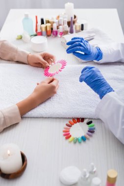 cropped view of woman holding palette of fake nails near manicurist and manicure supplies on blurred background clipart