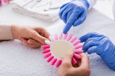 Manikürcünün, müşterinin elindeki sahte tırnak paletini parmağıyla göstermesinin kısmi görüntüsü