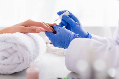 Lastik eldivenli manikürcünün ön plandaki bulanık hastaya manikür yaparken tırnak cilası sürerken kısmi görüntüsü