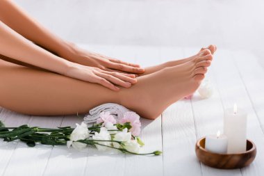 cropped view of woman with groomed hands and feet near white eustoma flowers and candles on white wooden surface clipart