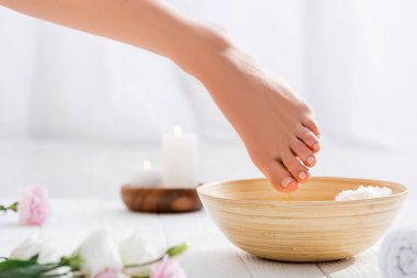 Pastel pembe tırnaklı kadın ayağı kasenin yanında ve beyaz yüzeyde çiçekler, bulanık ön plan