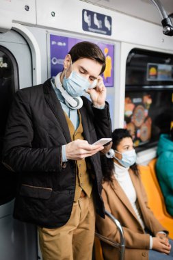 Tıbbi maskeli ve kulaklıklı bir adam metroda Afrikalı Amerikalı bir kadının yanında akıllı telefon kullanıyor. 