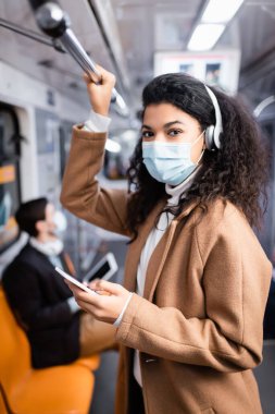 Kıvırcık Afrikalı Amerikan kadın tıbbi maske takıyor müzik dinliyor ve metroda akıllı telefon kullanıyor. 