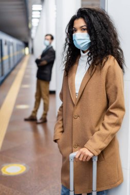 African american woman in medical mask standing with luggage in subway  clipart