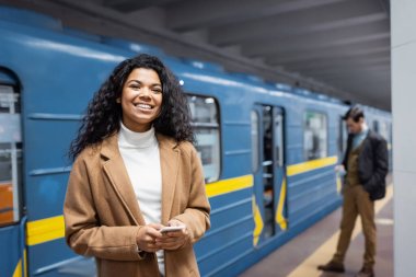 Akıllı telefonlu mutlu Afro-Amerikalı kadın metrodaki bulanık arka planda erkek ve arabanın yanında gülümsüyor. 