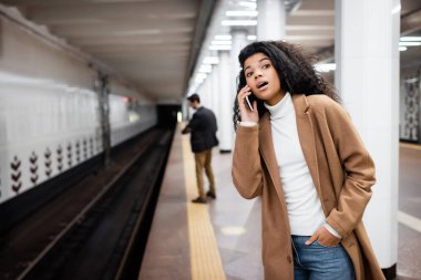 Şaşırmış Afrikalı Amerikalı kadın akıllı telefondan konuşuyor ve bulanık arka planda metroya bakıyor.