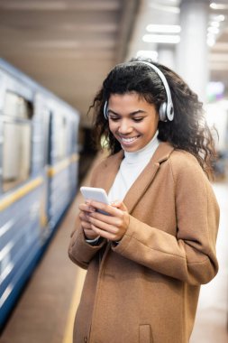 Kablosuz kulaklıklı gülümseyen Afrikalı Amerikalı kadın metroda müzik dinlerken akıllı telefon kullanıyor. 