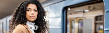 young african american woman in wireless headphones looking at camera in subway, banner clipart