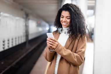 Kablosuz kulaklıklı gülümseyen Afrikalı Amerikalı kadın metroda akıllı telefon kullanıyor. 