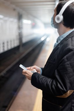Tıbbi maskeli ve kulaklıklı dövmeli adam metroda akıllı telefon tutuyor. 