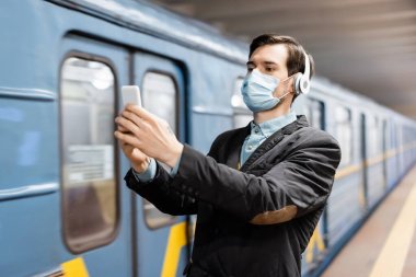 Tıbbi maskeli ve kablosuz kulaklıklı bir adam metroda akıllı telefondan selfie çekiyor. 