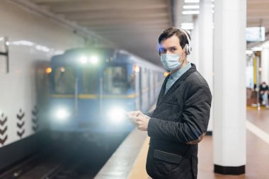 Tıbbi maskeli ve kablosuz kulaklıklı bir adam metroda akıllı telefon tutuyor. 