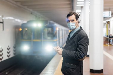 man in medical mask and wireless headphones holding smartphone in subway  clipart