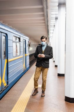 Tıbbi maskeli bir adam metro vagonunun yanında akıllı telefon tutuyor.