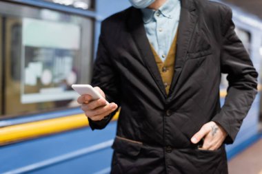 Tıbbi maskeli, akıllı telefon tutan ve elleri cebinde metro vagonunun yanında duran dövmeli adam görüntüsü.