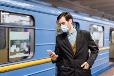Medikal maskeli adam akıllı telefon tutuyor ve elleri cebinde metro vagonunun yanında duruyor.