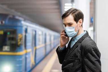 Tıbbi maskeli bir adam arka planda metro vagonunun yanında akıllı telefondan konuşuyor.