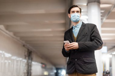 Tıbbi maskeli adam metro koridorunda ayakta ve akıllı telefonu tutuyor.