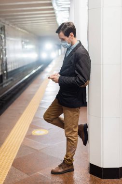 Tıbbi maskeli bir adam metro koridorunda ayakta duruyor ve akıllı telefonu tutuyor.
