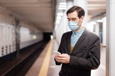 Tıbbi maskeli bir adam metroda ayakta duruyor ve akıllı telefon kullanıyor.