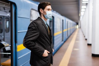 Tıbbi maskeli bir adam elleri ceplerinde metro vagonunun yanında duruyor.