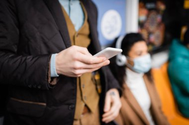 Metroda elinde akıllı telefon olan bir adam bulanık arka planda tıbbi maskeli Afrikalı Amerikalı bir kadının yanında.