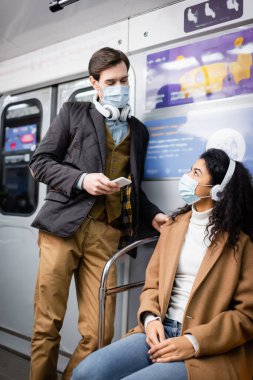 Kulaklıklı adam akıllı telefon tutuyor ve metrodaki tıbbi maskeli Afrikalı Amerikalı kadına bakıyor.