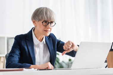 middle aged team leader in glasses showing thumb down and looking at laptop clipart