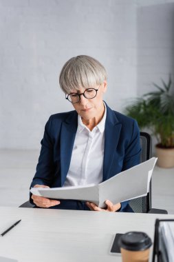 Gözlüklü orta yaşlı takım lideri bulanık önplanda kağıt bardağın yanında dokümanlara bakıyor.