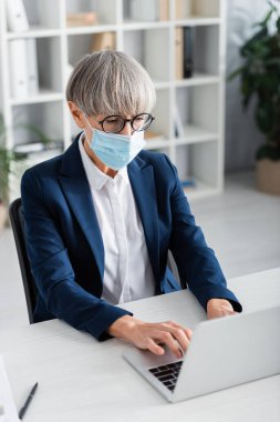 Gözlüklü ve tıbbi maskeli orta yaşlı takım lideri dizüstü bilgisayarda yazı yazıyor. 