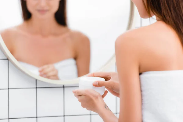 鏡の中のぼやけた反射の近くに美容クリームの容器を開く若い女性のクロップドビュー — ストック写真