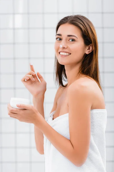 happy young woman with face cream on nose looking at camera in bathroom