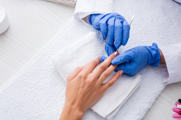 partial view of manicurist in latex gloves making manicure to woman with cuticle pusher